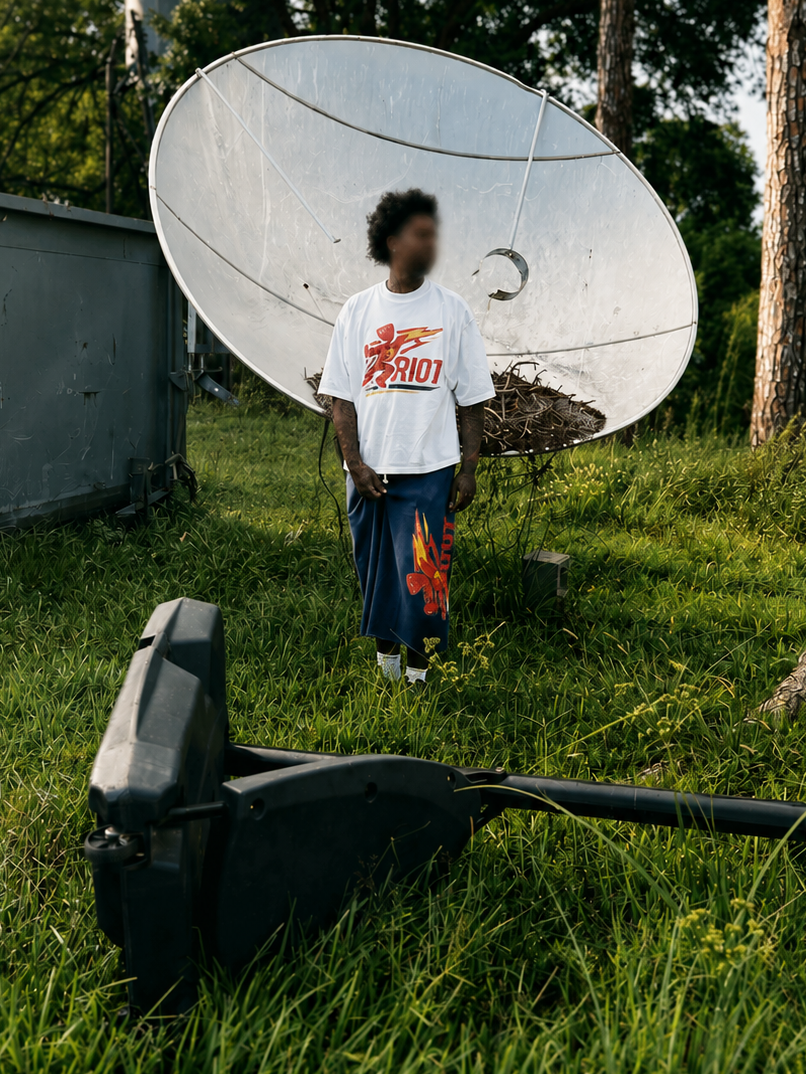 A person standing outdoors in a grassy area in front of a large white satellite dish, wearing a white t-shirt with a red and yellow graphic that reads 'I HOY' and dark blue pants with a red flame graphic.
